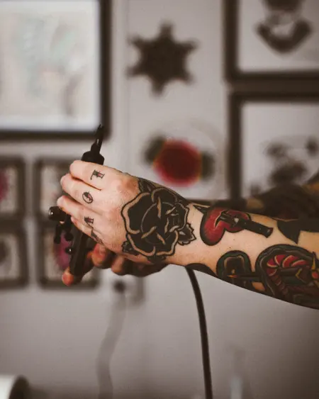 Traditional colour work up a forearm and a black rose on the hand, holding a tattoo machine.